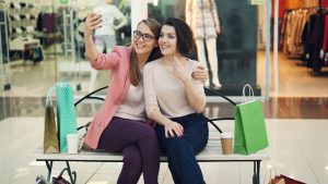 Amigas tirando selfie no shopping, sentadas no banco.